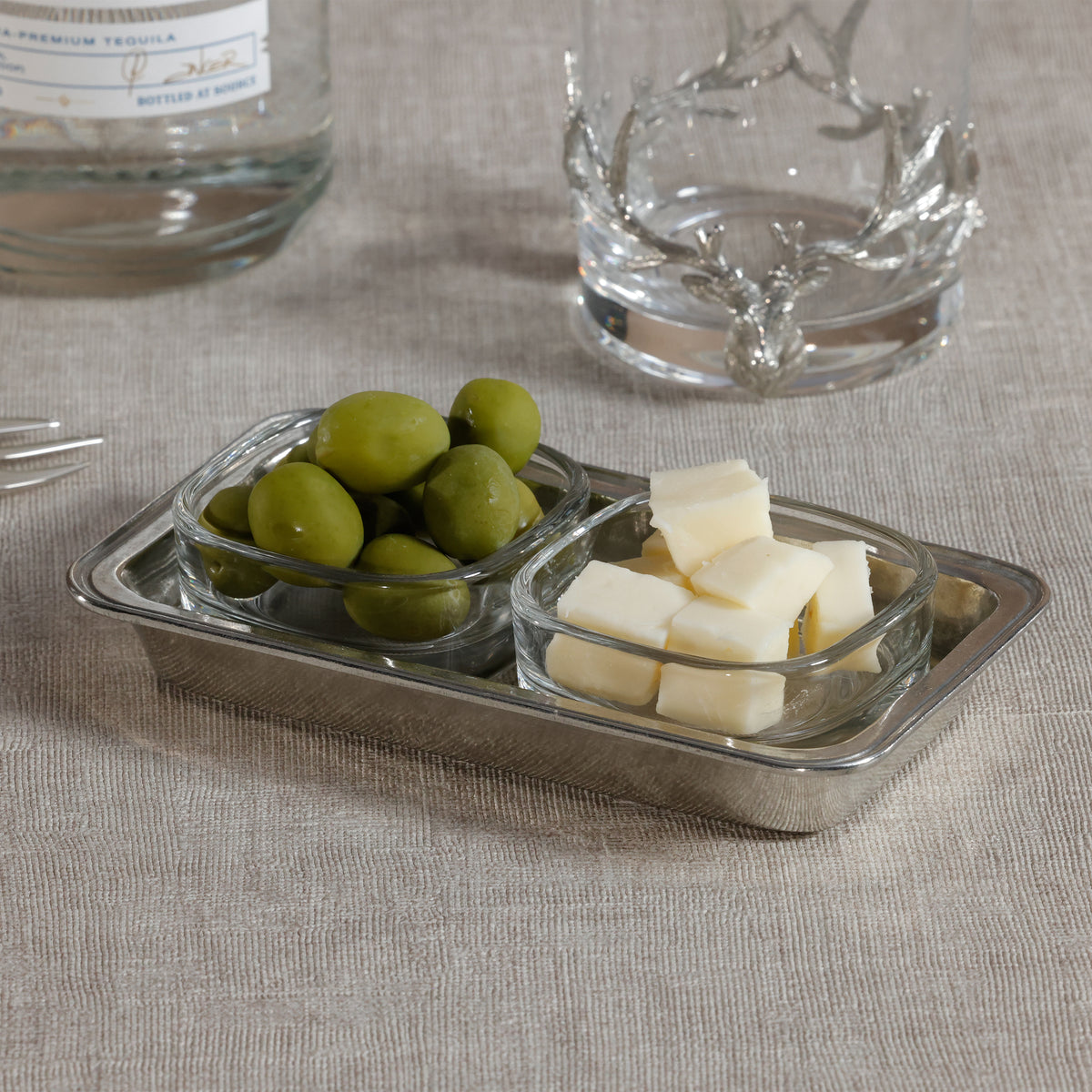 Small glass containers with olives and cheese on a textured surface with a bottle and glass in the background.